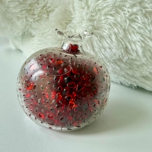 Pu&ograve; includere: Un oggetto decorativo a forma di melograno in vetro trasparente riempito di pezzi di vetro rossi e rosso scuro. La parte superiore ha un design a forma di corona. L'oggetto &egrave; su una superficie bianca.