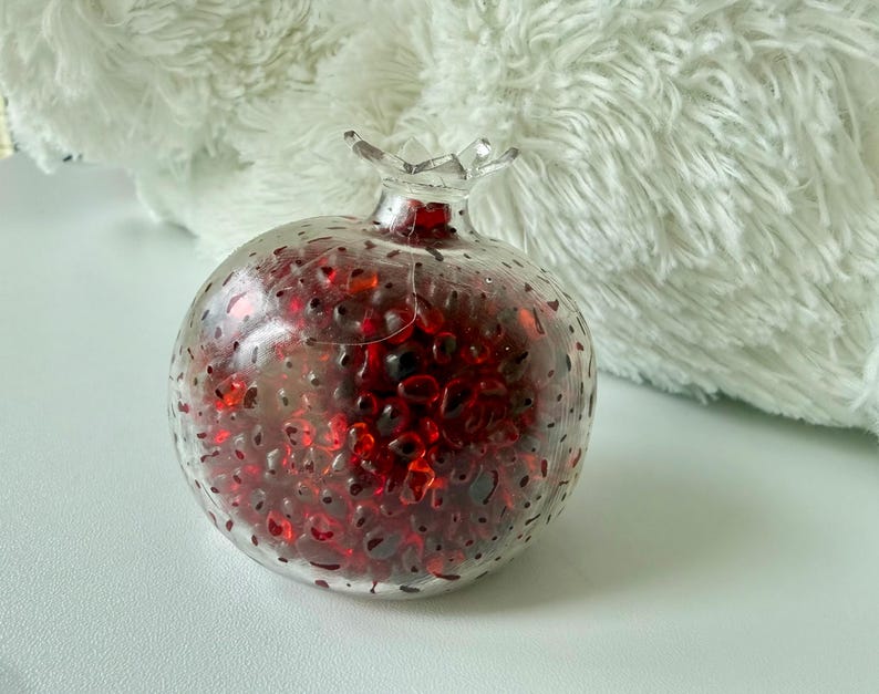 Pu&ograve; includere: Un oggetto decorativo a forma di melograno in vetro trasparente riempito di pezzi di vetro rossi e rosso scuro. La parte superiore ha un design a forma di corona. L'oggetto &egrave; su una superficie bianca.