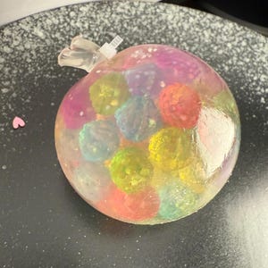 May include: A clear plastic sphere filled with colorful, translucent, round objects. The sphere has a small, clear plastic tube attached to the top.
