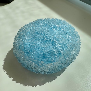 May include: A round, light blue decorative object with a textured, crystalline surface. The object is illuminated by sunlight, casting a shadow on the white surface below. The overall shape is spherical, with a rough, uneven texture.