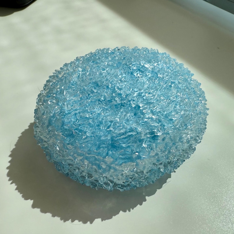 May include: A round, light blue decorative object with a textured, crystalline surface. The object is illuminated by sunlight, casting a shadow on the white surface below. The overall shape is spherical, with a rough, uneven texture.