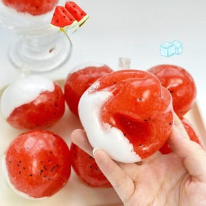 Puede incluir: Una colección de esferas de slime rojas y brillantes con pequeñas motas negras y una capa blanca opaca y gruesa en un lado. La textura parece gelatinosa y semitranslúcida, similar a un juguete sensorial con temática de fruta.
