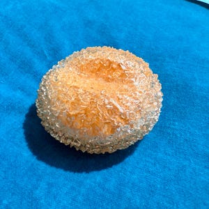 May include: A round, orange-colored object with a textured, crystalline exterior. The object is set against a blue background, creating a contrast in color and texture. The object appears to be a decorative item.