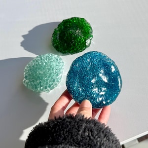 Op de afbeelding: Drie kleurrijke, getextureerde, glasachtige objecten in tinten groen, blauwgroen en blauw. Eén wordt vastgehouden door een hand met een zwarte, fuzzy mouw. De objecten hebben een bubbelachtige, organische vorm en worden op een wit oppervlak weergegeven.