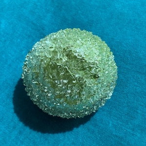 May include: A spherical, textured object with a light green hue. The surface has a bubbly, irregular pattern, creating a unique, organic appearance. The object is set against a teal background.