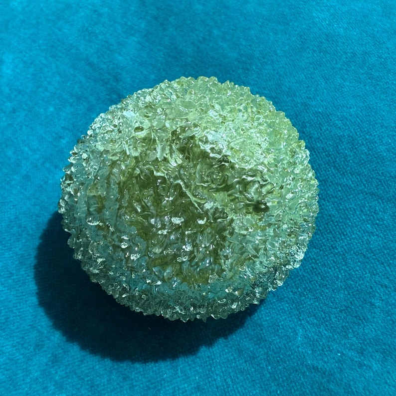 May include: A spherical, textured object with a light green hue. The surface has a bubbly, irregular pattern, creating a unique, organic appearance. The object is set against a teal background.