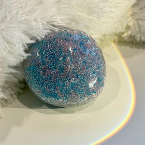 Könnte beinhalten: Ein abgerundeter, klarer Gegenstand, gefüllt mit kleinen, funkelnden blauen und lila Perlen. Das Objekt befindet sich auf einer weißen Oberfläche, mit einer flauschigen weißen Textur im Hintergrund. Ein Regenbogenbogen ist auf der rechten Seite sichtbar.