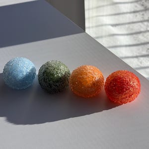 May include: Four textured spheres in a row, in shades of blue, green, orange, and red. The spheres are on a white surface, with shadows cast by sunlight. The spheres are approximately 5 cm in diameter.
