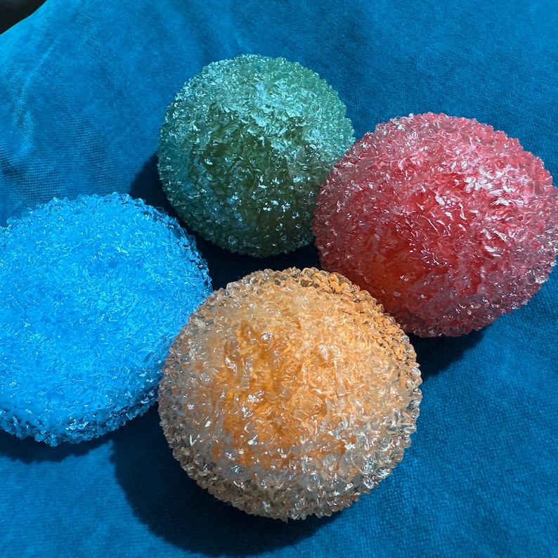 May include: Four colorful, textured spheres in blue, green, orange, and red. The spheres have a crystalline appearance and are arranged on a blue fabric background. The spheres are round and appear to be made of glass or a similar material.