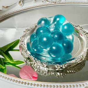May include: A cluster of blue, translucent, bubble-like spheres arranged in a silver, ornate dish. The dish is on a white, mirrored surface.
