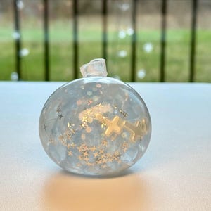 以下が含まれることがあります： 明るい青色の液体、金色の星、金色の十字架が詰められた、透明で丸いオーナメント。オーナメントには小さな透明な上部があります。背景は、緑色の屋外シーンのぼやけたビューです。