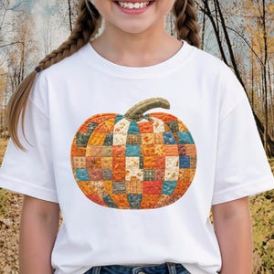 May include: A white short-sleeved t-shirt featuring a large, central graphic of a pumpkin constructed from a patchwork of various quilted fabric squares in shades of orange, blue, yellow, and brown, topped with a textured green stem.