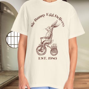 以下が含まれることがあります： クリーム色のTシャツ。ヴィンテージ風のイラストで、自転車に乗ったウサギが卵を運んでいます。「Sir Bunny Egg Delivery」と「EST. 1980」の文字が茶色で書かれています。半袖でクルーネックのシャツです。