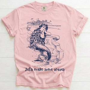 May include: A light pink t-shirt featuring a navy blue illustration of a mermaid leaning on a railing. The mermaid has long hair and a fishtail. The text "Let's make some waves" is printed below the illustration.