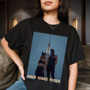 May include: A black t-shirt featuring a graphic print of two individuals standing before a tall skyscraper at dusk. The person on the left wears cat ears, and the person on the right wears a cap. The text AMOR SOBRE PODER is printed in gold.