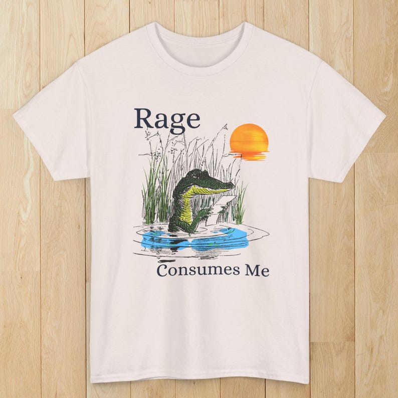 Puede incluir: Una camiseta blanca de manga corta con un gr&aacute;fico de un caim&aacute;n verde en el agua rodeado de hierba alta y un sol poniente. El texto "Rage" aparece sobre el caim&aacute;n y "Consumes Me" est&aacute; impreso debajo de la ilustraci&oacute;n del agua.