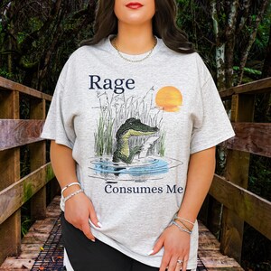 Puede incluir: Una camiseta gris claro de manga corta con un gr&aacute;fico de un caim&aacute;n en un pantano con hierba alta y un sol poniente. El texto "Rage" est&aacute; impreso sobre el caim&aacute;n y el texto "Consumes Me" est&aacute; impreso debajo del gr&aacute;fico en azul oscuro.