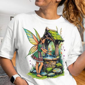 May include: A white t-shirt featuring a graphic illustration of a fairy with large, colorful wings standing beside a stone well. The fairy wears a green dress with intricate patterns, and the scene includes mossy stones and wildflowers.