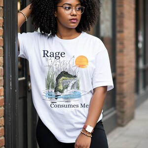 Puede incluir: Una camiseta blanca de manga corta con un estampado gr&aacute;fico de un caim&aacute;n en el agua con hierba alta y una puesta de sol. El texto "Rage" est&aacute; impreso sobre el caim&aacute;n y el texto "Consumes Me" aparece en azul oscuro bajo el gr&aacute;fico.