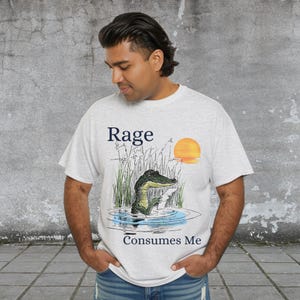 Puede incluir: Una camiseta gris claro de manga corta con un gr&aacute;fico de un caim&aacute;n verde en el agua rodeado de hierba alta y un sol naranja. El texto "Rage" est&aacute; impreso sobre el caim&aacute;n y el texto "Consumes Me" est&aacute; impreso debajo del gr&aacute;fico.