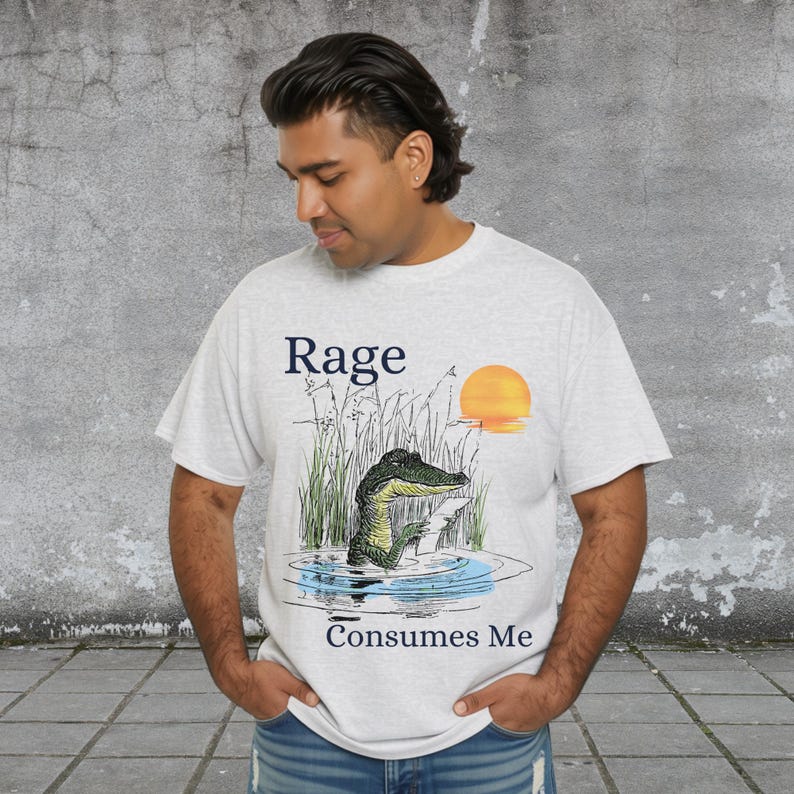 Puede incluir: Una camiseta gris claro de manga corta con un gr&aacute;fico de un caim&aacute;n verde en el agua rodeado de hierba alta y un sol naranja. El texto "Rage" est&aacute; impreso sobre el caim&aacute;n y el texto "Consumes Me" est&aacute; impreso debajo del gr&aacute;fico.