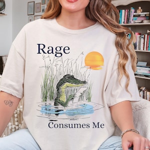 Puede incluir: Una camiseta de color crema con una ilustraci&oacute;n gr&aacute;fica de un caim&aacute;n en un estanque pantanoso al atardecer. El texto "Rage" aparece sobre el caim&aacute;n y "Consumes Me" est&aacute; impreso debajo. El caim&aacute;n sostiene un peque&ntilde;o papel de color blanco.