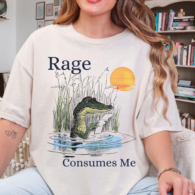 Puede incluir: Una camiseta de color crema con una ilustraci&oacute;n gr&aacute;fica de un caim&aacute;n en un estanque pantanoso al atardecer. El texto "Rage" aparece sobre el caim&aacute;n y "Consumes Me" est&aacute; impreso debajo. El caim&aacute;n sostiene un peque&ntilde;o papel de color blanco.