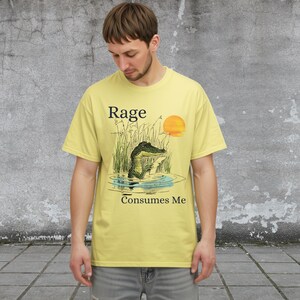 Puede incluir: Una camiseta de color amarillo p&aacute;lido con una ilustraci&oacute;n gr&aacute;fica de un caim&aacute;n en un pantano con hierba alta y un sol poniente. El texto "Rage" est&aacute; impreso sobre el caim&aacute;n y "Consumes Me" est&aacute; impreso debajo de la ilustraci&oacute;n.