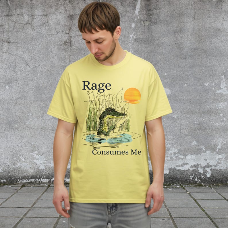 Puede incluir: Una camiseta de color amarillo p&aacute;lido con una ilustraci&oacute;n gr&aacute;fica de un caim&aacute;n en un pantano con hierba alta y un sol poniente. El texto "Rage" est&aacute; impreso sobre el caim&aacute;n y "Consumes Me" est&aacute; impreso debajo de la ilustraci&oacute;n.