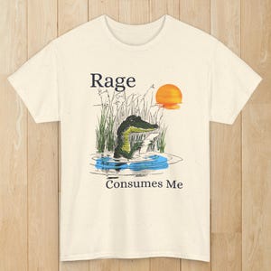Puede incluir: Una camiseta de color crema con una ilustraci&oacute;n gr&aacute;fica de un caim&aacute;n en el agua rodeado de hierba alta y una puesta de sol. El texto "Rage" aparece sobre el caim&aacute;n y "Consumes Me" est&aacute; impreso debajo del agua en letras negras.