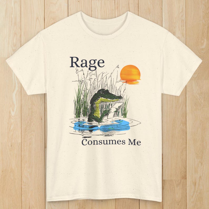 Puede incluir: Una camiseta de color crema con una ilustraci&oacute;n gr&aacute;fica de un caim&aacute;n en el agua rodeado de hierba alta y una puesta de sol. El texto "Rage" aparece sobre el caim&aacute;n y "Consumes Me" est&aacute; impreso debajo del agua en letras negras.