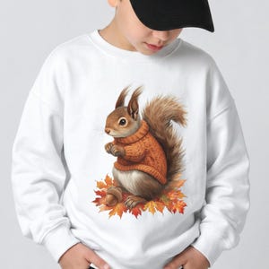 May include: A white long-sleeve sweatshirt featuring a graphic print of a brown squirrel wearing an orange knit sweater. The squirrel is sitting on a pile of colorful autumn leaves and holding a brown acorn. The design is centered on the chest.