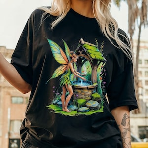 May include: Black t-shirt featuring a colorful illustration of a fairy near a well. The fairy has green and yellow wings and is wearing a green outfit. The well is made of stone and has a wooden roof. The background includes flowers and greenery.