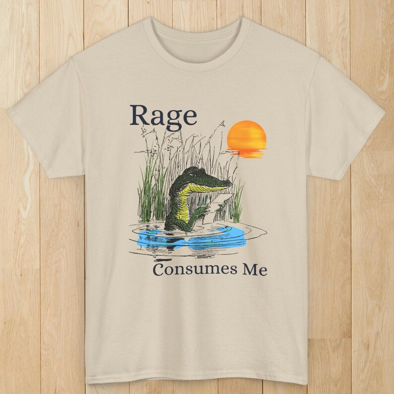 Puede incluir: Una camiseta beige de manga corta con un gr&aacute;fico de un caim&aacute;n en el agua rodeado de juncos. El caim&aacute;n sostiene un papel y se ve un sol naranja brillante. El texto "Rage" aparece arriba del gr&aacute;fico y "Consumes Me" est&aacute; debajo.