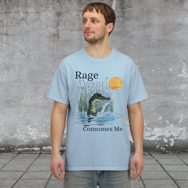 Puede incluir: Una camiseta de manga corta de color azul claro con un gr&aacute;fico de un caim&aacute;n en el agua rodeado de hierba alta y un sol amarillo. El texto "Rage" est&aacute; impreso sobre el caim&aacute;n y el texto "Consumes Me" est&aacute; impreso debajo del gr&aacute;fico.