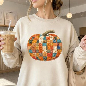 Op de afbeelding: Een crèmekleurig sweatshirt met ronde hals en een grote, centrale afbeelding van een pompoen gemaakt van een patchwork van diverse kleurrijke stoffen vierkantjes in tinten oranje, blauw, geel en bruin, met een gestructureerde groene steel.
