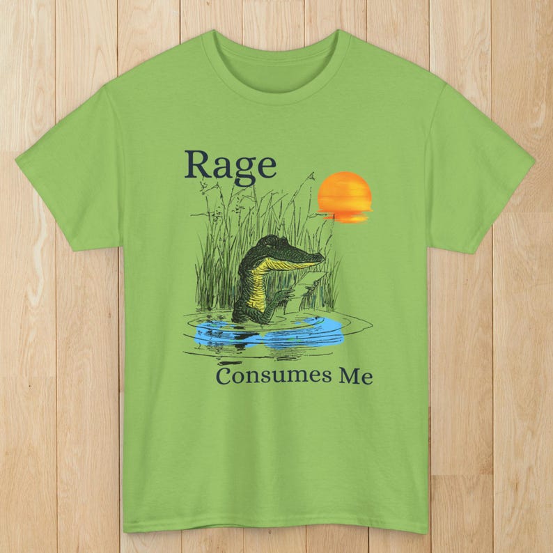 Puede incluir: Una camiseta de manga corta de color verde lima con una ilustraci&oacute;n gr&aacute;fica de un caim&aacute;n en un pantano con hierba alta y un sol poniente. El texto "Rage" est&aacute; impreso sobre el caim&aacute;n y "Consumes Me" est&aacute; impreso debajo de la ilustraci&oacute;n.