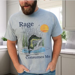 Puede incluir: Una camiseta azul claro con un gr&aacute;fico de un caim&aacute;n en el agua rodeado de hierba alta y un sol amarillo. El texto "Rage" est&aacute; impreso sobre el caim&aacute;n y el texto "Consumes Me" est&aacute; impreso debajo del gr&aacute;fico en color azul oscuro.
