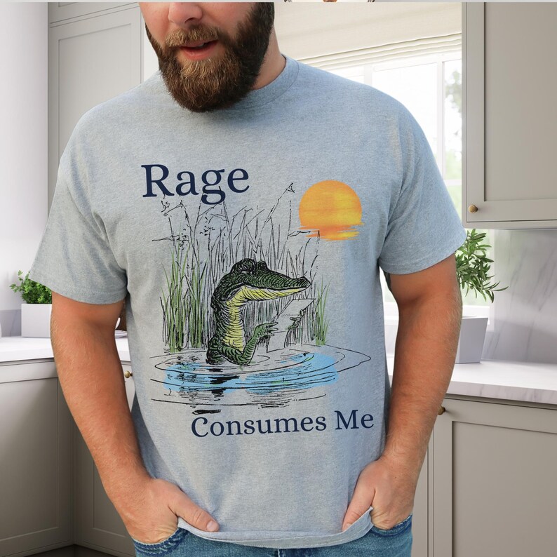 Puede incluir: Una camiseta azul claro con un gr&aacute;fico de un caim&aacute;n en el agua rodeado de hierba alta y un sol amarillo. El texto "Rage" est&aacute; impreso sobre el caim&aacute;n y el texto "Consumes Me" est&aacute; impreso debajo del gr&aacute;fico en color azul oscuro.
