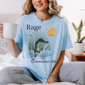 Puede incluir: Una camiseta azul claro con un gr&aacute;fico de un caim&aacute;n en el agua rodeado de hierba alta y un sol amarillo. El texto "Rage" est&aacute; impreso sobre el caim&aacute;n y "Consumes Me" est&aacute; impreso debajo del gr&aacute;fico en letras de color azul oscuro.