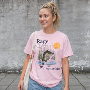 Puede incluir: Una camiseta de manga corta de color rosa claro con una ilustraci&oacute;n gr&aacute;fica de un caim&aacute;n en un pantano con una puesta de sol. El texto "Rage" est&aacute; impreso sobre el caim&aacute;n y "Consumes Me" debajo en letras de color azul oscuro.