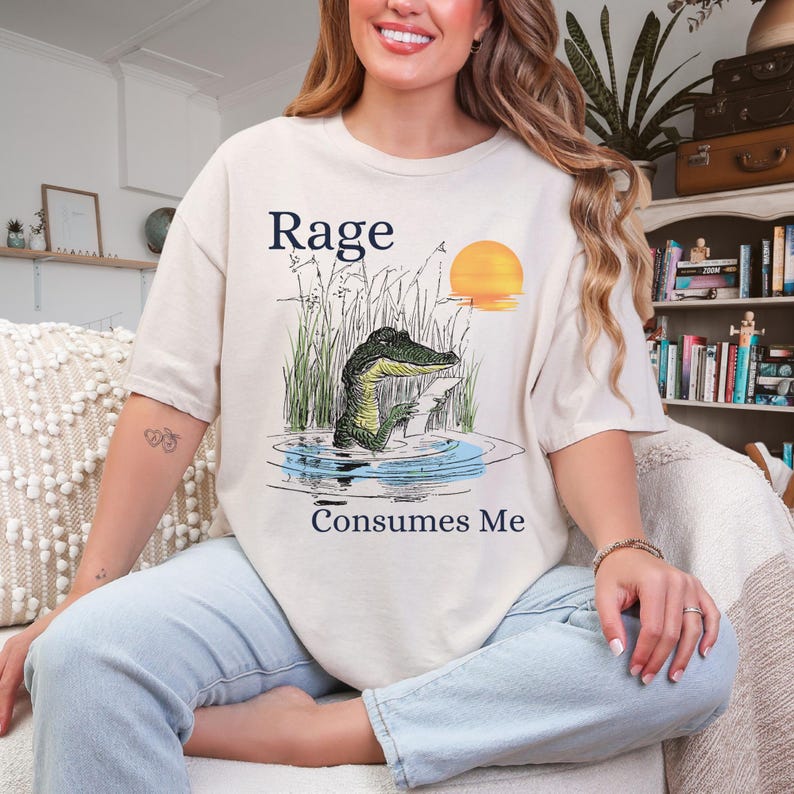 Puede incluir: Una camiseta de manga corta de color crema con una ilustraci&oacute;n gr&aacute;fica de un caim&aacute;n en el agua rodeado de hierba alta y un sol amarillo. El texto "Rage" est&aacute; impreso sobre el caim&aacute;n y "Consumes Me" est&aacute; impreso debajo del gr&aacute;fico.
