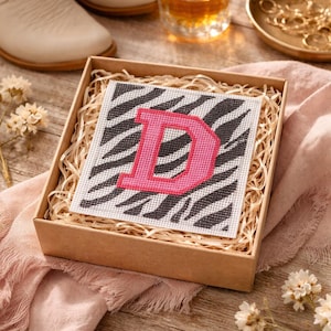 May include: A decorative box with a canvas featuring a pink letter "D" on a black and white zebra print. The box is filled with straw and sits on a wooden surface, with a glass of amber liquid and a pair of white boots in the background.