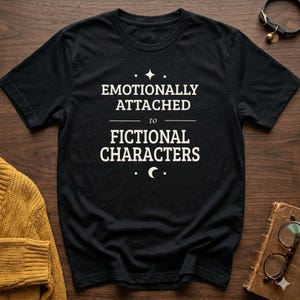 May include: A black short-sleeved t-shirt featuring the white text "EMOTIONALLY ATTACHED to FICTIONAL CHARACTERS" centred on the chest. The design includes a small four-pointed star above the text and a crescent moon symbol below the text.