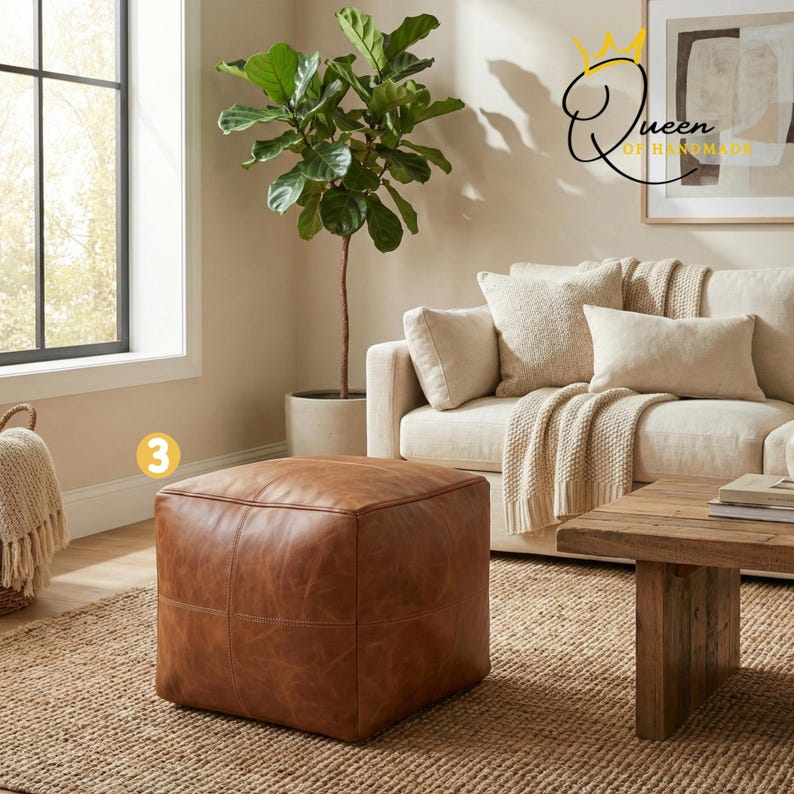 May include: A square, brown leather ottoman with stitched detailing. The ottoman is placed on a woven jute rug in a living room setting. A sofa with cream-colored cushions and a wooden coffee table are also visible.