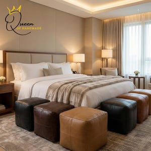 May include: A luxurious bedroom setting featuring a large bed with white linens and a plush, neutral-toned throw blanket. Six leather cube ottomans in shades of black, brown, and tan are arranged in front of the bed. The room has warm lighting and a neutral color palette.