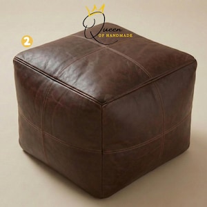 May include: A square, brown leather ottoman with stitched detailing. The ottoman is a cube shape and has a smooth, slightly distressed appearance. The stitching is a lighter brown color, creating a subtle contrast. The product is from Queen of Handmade.