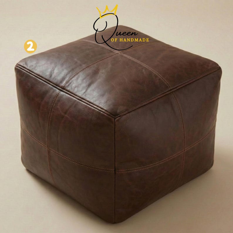 May include: A square, brown leather ottoman with stitched detailing. The ottoman is a cube shape and has a smooth, slightly distressed appearance. The stitching is a lighter brown color, creating a subtle contrast. The product is from Queen of Handmade.