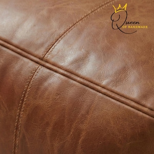 May include: Close-up of a brown leather surface with visible stitching. The leather has a textured appearance and a warm, inviting color. The image also includes the text "Queen of Handmade" with a crown logo.