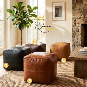 May include: Four leather ottomans in varying shades of brown and black are arranged on a woven rug. The ottomans are square-shaped with stitched detailing. The room features a large plant, a wooden coffee table, and a stone fireplace.
